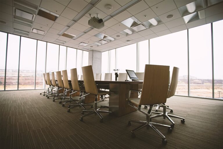 Empty conference room with a long table, swivel chairs, and large windows providing natural light. Ceiling features lights and ventilation panels.