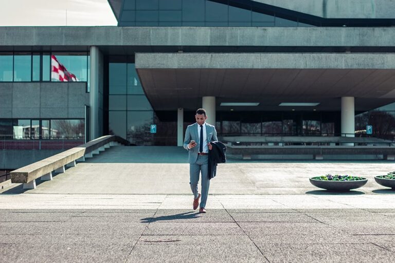 A man in a suit walks outside a modern building while looking at his phone. A flag and potted plants are visible in the background.