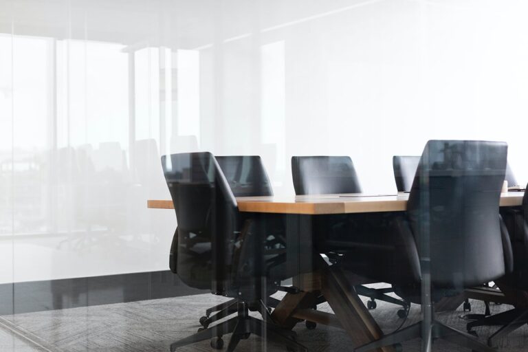 Empty conference room with a long wooden table and eight black chairs. Large windows allow natural light to fill the space, and the room is set for a meeting.