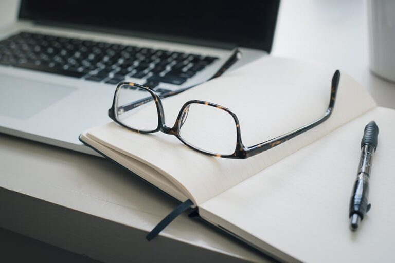 Glasses resting on an open notebook with a pen beside it, next to a laptop on a desk.