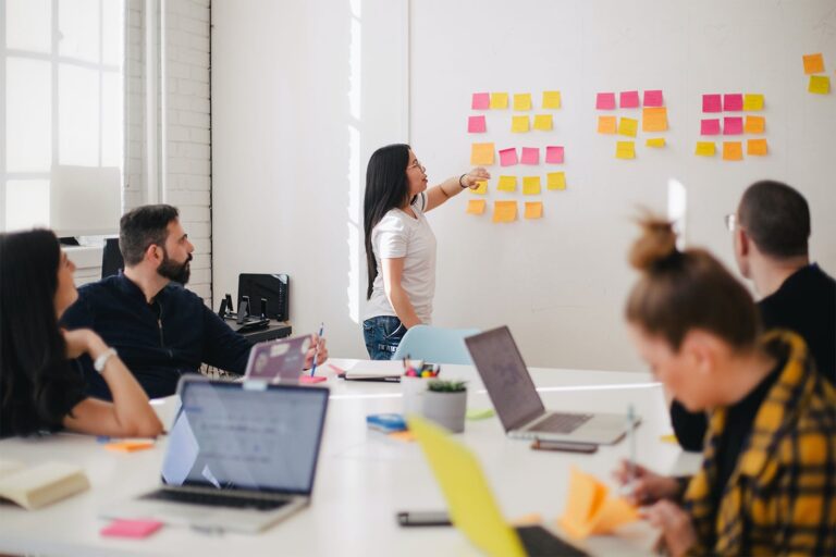 Person presenting sticky notes on a whiteboard to a group of people in a bright office, with laptops on the table.